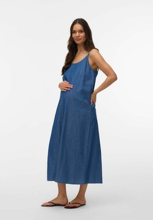 Gravid kvinne med langt mørkt hår iført ermeløs blå denim maxi-kjole og svarte flip-flops, som forsiktig holder magen, stående mot hvit bakgrunn.