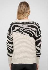 Chunky stickad tröja med ett svart och vitt zebramönster på baksidan, ribbade ärmslut och en neutral beige kropp med horisontella ränder.