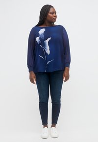 Γυναίκα με πλεξούδες που φοράει μπλούζα ναυτικού με λευκά λουλούδια, σκούρα στενά τζιν και λευκά αθλητικά παπούτσια, στέκεται με χαλαρά χέρια.