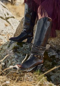 Cizme negre din piele până la genunchi, cu vârfuri ascuțite, decorate cu accente metalice și curele suprapuse. Toc robust și textură netedă.