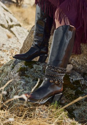 Czarne skórzane buty za kolano z ostrymi czubkami, ozdobione metalicznymi akcentami i warstwowymi paskami. Gruby obcas i gładka faktura.