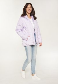 Jachetă pufoasă de culoare lavandă, cu glugă, design matlasat, cu fermoar frontal și buzunare laterale, asortată cu blugi ușor colorați în albastru deschis și adidași albi.