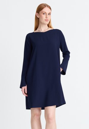 Frau mit glatten roten Haaren, die ein langärmeliges, marineblaues A-Linien-Kleid mit einer silbernen Halskette auf hellem Hintergrund trägt.