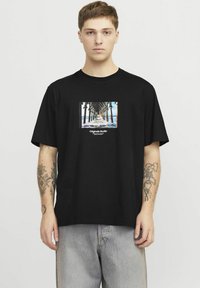 Juodas medvilninis marškinėliai su grafiniu piešiniu, vaizduojančiu prieplauką ir vandenyną, žemiau vaizdo esant tekstui "Originals Studio". Trumpomis rankovėmis, laisvas kirpimas.