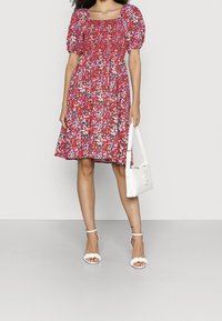 Robe florale rouge avec des manches bouffantes courtes, un décolleté carré et un corps élastique, associée à un sac blanc et des sandales à lanières.