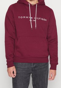 Felpa maroon realizzata in tessuto misto cotone, con una tasca frontale, cordino bianco e testo del logo sulla parte anteriore.