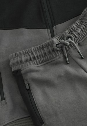 Pantaloni jogger grigi con vita regolabile tramite cordoncino, dettagli neri e tasche con zip, realizzati in materiale morbido e leggero.