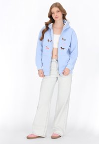 Hellblauer Kapuzenpullover mit Reißverschluss und Tierstickerei, kombiniert mit weißem Croptop und hellblauen, weit geschnittenen Jeans. Auffällige Kapuze mit Spitzenbesatz.