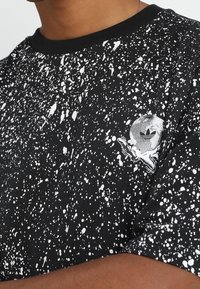 Černé tričko s bílou malbou ve tvaru skvrn a malým logem Adidas na levé části hrudi, které má na sobě osoba se středním hnědým tónem pleti.