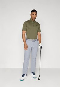 Olívazöld galléros póló, szürke nadrág és fehér golfcipő. A modell golfütőt tart, és fehér kesztyűt visel. Egyszerű, testhezálló kialakítás.