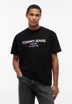 Νεαρός άνδρας που φοράει μαύρο t-shirt "Tommy Jeans" με πολύχρωμο κείμενο και σκούρο τζιν, στέκεται με το ένα χέρι στην τσέπη μπροστά σε λευκό φόντο.
