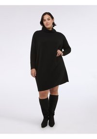 Vestito nero in maglia con collo a lupetto, maniche lunghe e orlo asimmetrico. Indossato con stivali neri in camoscio al ginocchio.