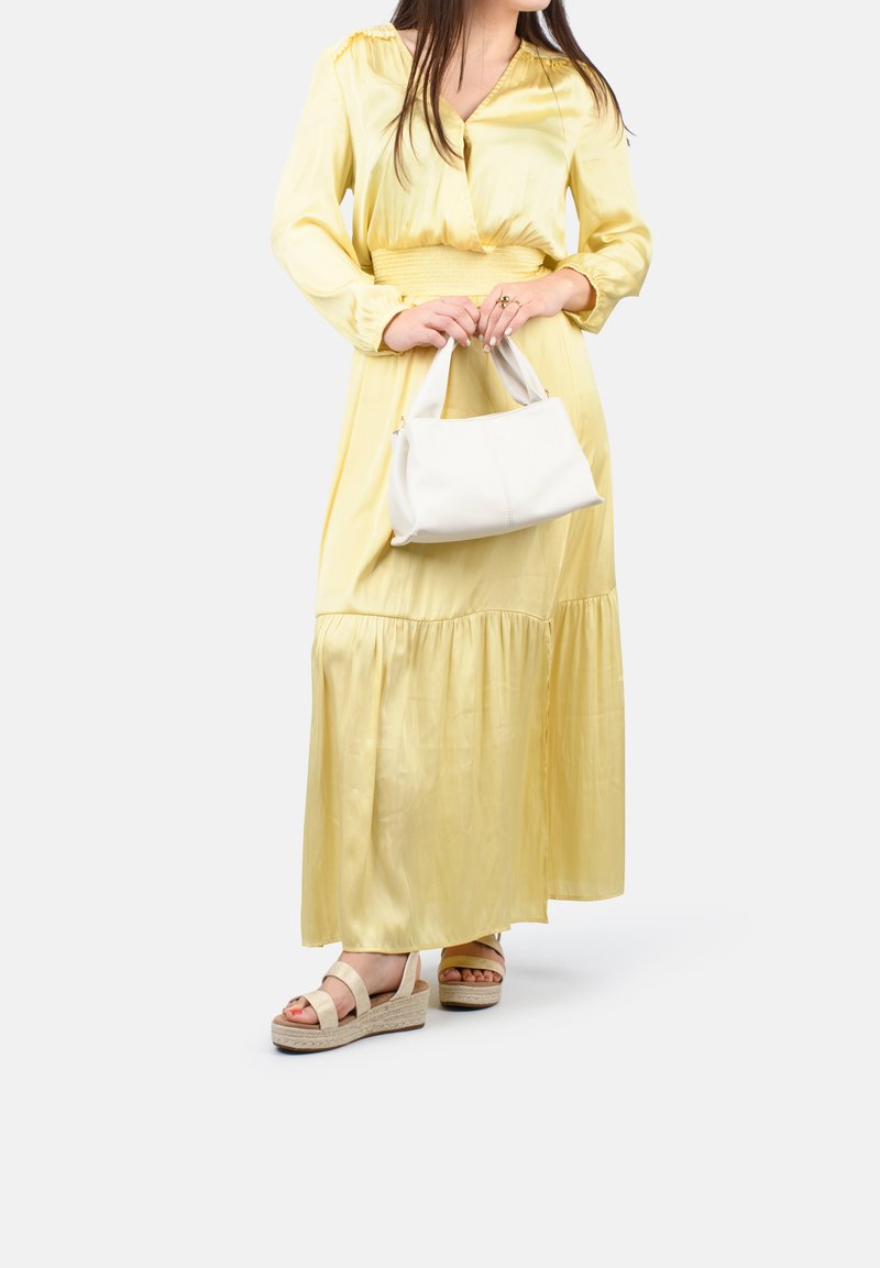 Long yellow satin dress with a cinched waist, paired with beige wedge sandals and a white handbag. Smooth texture and flowing design.
