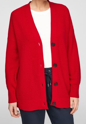 Cardigan rouge en maille avec boutons noirs porté sur un haut blanc et un pantalon bleu marine à cordon, visible du cou aux hanches.