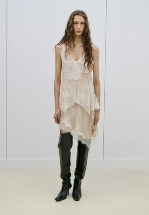 Mujer con cabello largo y rizado, vestida con un vestido de encaje beige asimétrico y botas negras de tacón alto hasta la rodilla, sobre un fondo blanco liso.