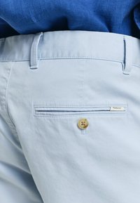 Světle modré kalhoty vyrobené z měkké tkaniny s jednou zadní kapsou, mají dřevěný knoflík a bílou štítek s nápisem "GANT".
