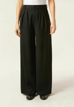 Persona che indossa pantaloni neri a gamba larga con vita alta e pieghe, abbinati a scarpe nere e top bianco senza maniche.
