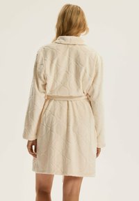 Kremowy szlafrok o pikowanym wzorze, z długimi rękawami, kołnierzem typu szal oraz paskiem w talii. Miękka, teksturowana tkanina.