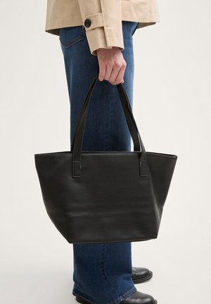 Schwarze Echtleder-Shopper-Tasche mit zwei kurzen Griffen, rechteckiger Form, strukturierter Oberfläche und flachem Boden. Wird von einer Person mit blauen Jeans getragen.