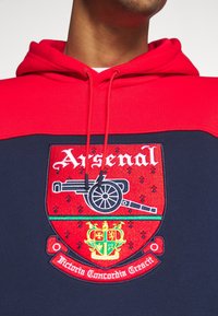 Röd och marinblå hoodie med ett broderat Arsenal-emblem med en kanon, gröna detaljer och texten "Victoria Concordia Crescet".