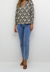 Femme portant un blouse à motifs géométriques beige et noir, un jean bleu clair et des sandales à talons ouvertes marron, se tenant contre un mur blanc.