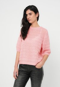 Ροζ πλεκτό πουλόβερ με κοντά μανίκια, με υφασμένο μοτίβο και στρογγυλή λαιμόκοψη, συνδυασμένο με σκουρόχρωμο, ελαφρώς υφασμένο παντελόνι.