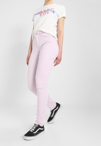 Persona vestida con una camiseta blanca que dice "Maybe Baby", jeans skinny rosa claro y zapatillas negras Vans, de pie contra un fondo claro y simple.