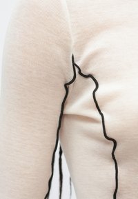 Top cu mâneci lungi din țesătură bej deschis cu dungi, având cusături contrastante negre de-a lungul cusăturilor și accente texturate.