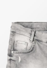Jeans de mezclilla gris con un diseño desgastado, que cuentan con un bolsillo frontal, cierre de botón y acentos de costura. Textura suave con un ligero brillo.