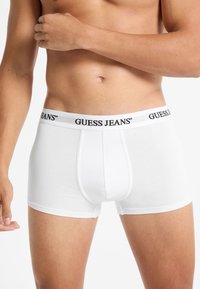 Boxer da uomo bianchi con logo "GUESS JEANS" nero sull'elastico in vita. Tessuto liscio, forma aderente e lunghezza fino a metà coscia.