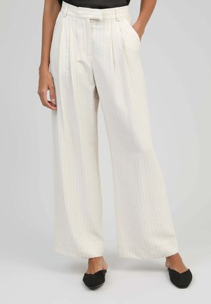 Pantalon large crème à fines rayures, taille haute avec passants de ceinture, porté avec des chaussures noires à bout pointu, mains dans les poches.