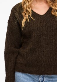 Maglione marrone lavorato a maglia con scollo a V, caratterizzato da un motivo testurizzato e maniche lunghe, abbinato a jeans in denim azzurro chiaro.