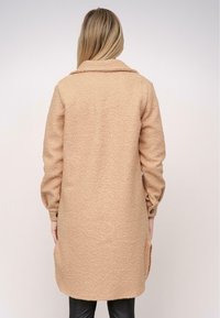 Beige, texturovaný kabát s límečkem, dlouhými rukávy a bočními kapsami; má mírně oversized střih a délku po kolena.