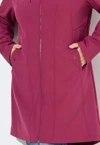 Maroon softshell coat with a full zipper, side pockets, and a fitted design. Smooth texture, no visible patterns or embellishments.