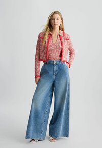 Giacca cropped in tweed rosso con colletto e dettagli con bottoni abbinata a jeans a gamba larga di un blu chiaro. La modella indossa sandali bianchi con punta aperta.