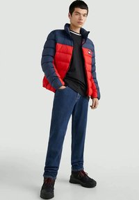 Νεαρός άνδρας στέκεται με τα χέρια στις τσέπες, φορώντας ένα κόκκινο και ναυτικό μπουφάν, μαύρο πουκάμισο, μπλε τζιν και μαύρες μπότες με κόκκινες κορδόνες.