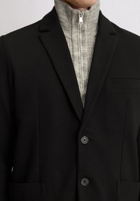 Schwarzes Blazer mit Reverskragen, zwei schwarzen Knöpfen und einer Brusttasche. Darunter ein hellgrauer, gerippter Rollkragenpullover mit einem Frontreißverschluss.
