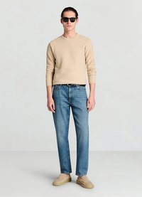Beiger, gerippter Pullover, hellblaue Jeans mit geradem Bein, beige Slip-on-Schuhe und schwarze Sonnenbrille. Einfaches, lässiges Outfit mit einer neutralen Farbpalette.