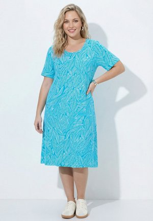 Smilende kvinde med blondt hår iført en blå knælængde kjole med hvide bladmønstre og hvide sneakers, poserende med den ene hånd på hoften.