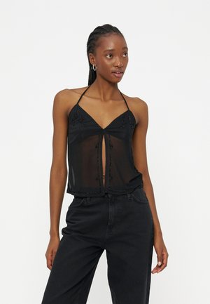 Νεαρή γυναίκα με διαφανές μαύρο τοπ halter με διακοσμήσεις από χάντρες και μαύρο ψηλόμεσο παντελόνι, ποζάροντας μπροστά σε απλό φόντο.