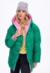 Zielona i różowa pikowana kurtka z kapturem, zapinana na zatrzaski, wykonana z błyszczącego materiału. Do tego szary dziany beret i żółta bluzka.