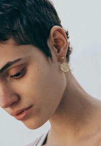 Guldörhängen med en rund skiva med ett upphöjt mönster. Ytan verkar slät, med en polerad finish. Små stickbackar.