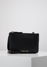 Czarna skórzana kopertówka o gładkiej fakturze, z złotym logo, zamkiem błyskawicznym i odpinanym paskiem. Kompaktowy, prostokątny kształt.