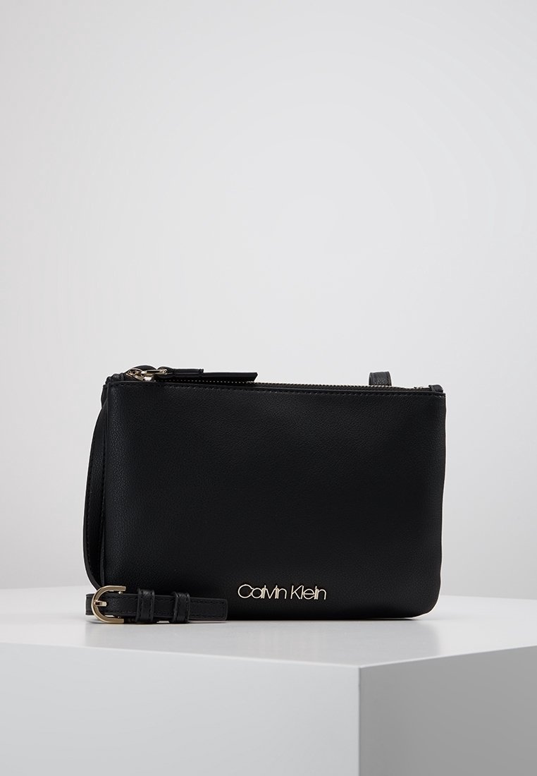 Czarna skórzana kopertówka o gładkiej fakturze, z złotym logo, zamkiem błyskawicznym i odpinanym paskiem. Kompaktowy, prostokątny kształt.