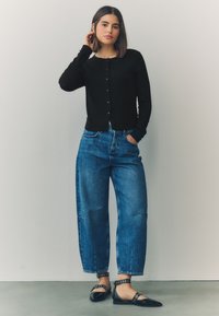 Black long-sleeve cardigan with buttons, paired with loose blue denim jeans and black flats featuring buckle accents. Simple, modern silhouette.