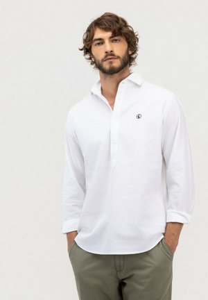 Hombre con cabello rizado y barba, vistiendo una camisa blanca abotonada con las mangas remangadas y pantalones verdes con las manos en los bolsillos.