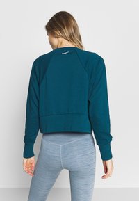 Sweat-shirt court bleu sarcelle avec col rond, manches raglan et un logo Nike discret dans le dos, associé à un legging gris clair.