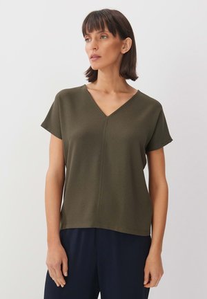 Top vert olive à col en V avec manches courtes, en tissu texturé, et une couture centrale. Associé à un pantalon marine, montrant une coupe décontractée.