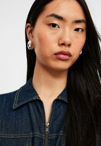 Tumman denim-takki, jossa on vetoketju edessä ja terävä kaulus. Suuret läpinäkyvät ja metalliset korvakorut, sileä ihon pinta.