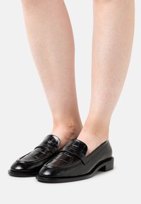 Mocasines de charol negro con un patrón texturizado de cocodrilo, tacón bajo y abertura en la parte posterior para facilitar su uso.